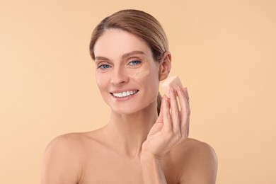 Woman blending foundation on face with makeup sponge against beige background