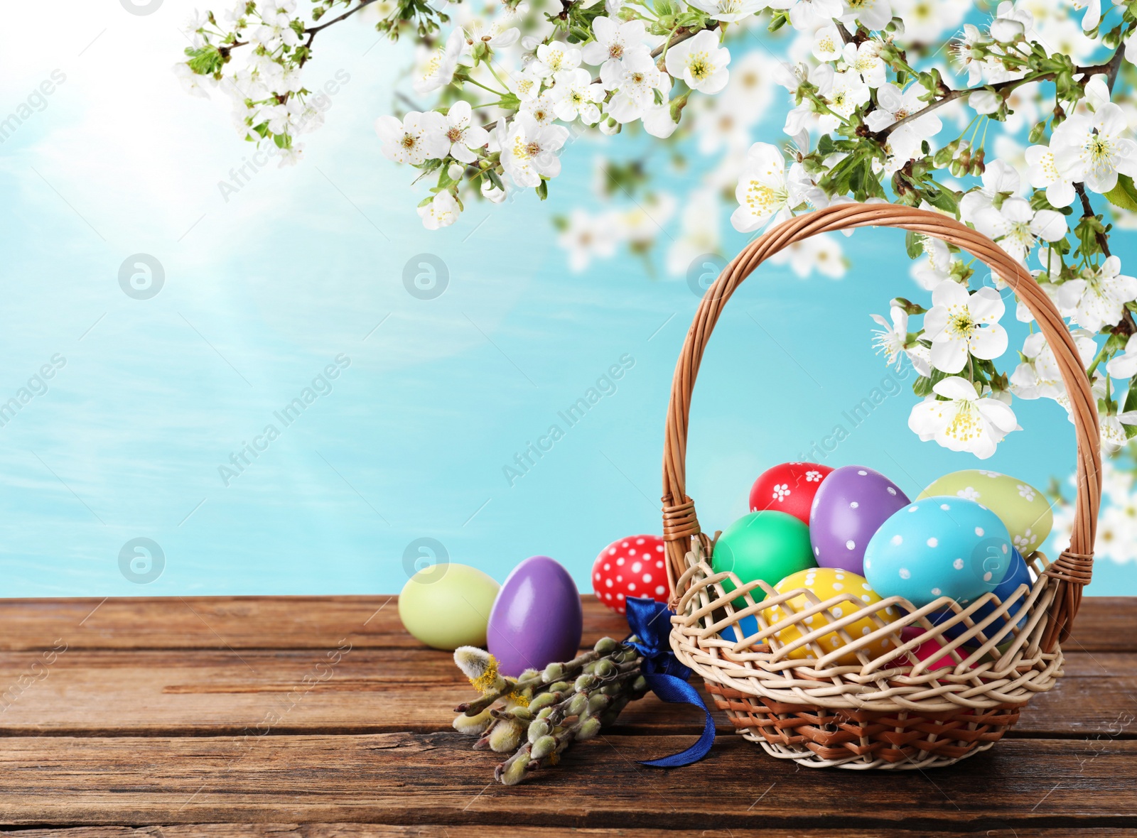 Image of Colorful Easter eggs in wicker basket and willow branches on wooden table outdoors, space for text