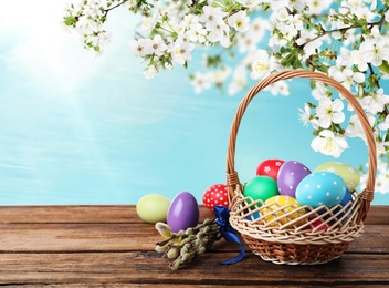 Image of Colorful Easter eggs in wicker basket and willow branches on wooden table outdoors, space for text