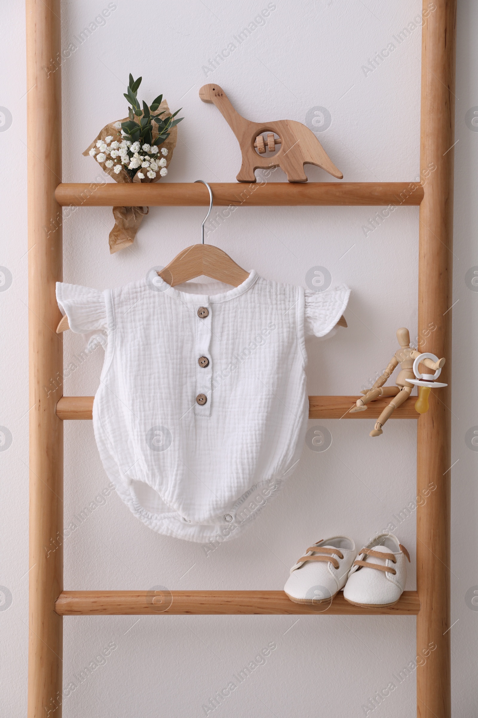 Photo of Baby bodysuit, shoes, toys and small bouquet on ladder near white wall