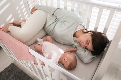 Tired young mother sleeping in crib with her baby at home, above view