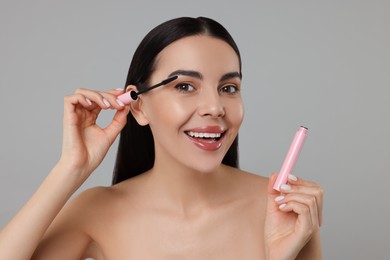 Beautiful young woman applying mascara on grey background