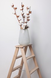 Photo of Cotton branches with fluffy flowers in vase on wooden ladder near white wall