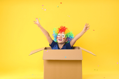 Photo of Little boy in clown wig sitting inside of cardboard box under confetti shower on yellow background. April fool's day
