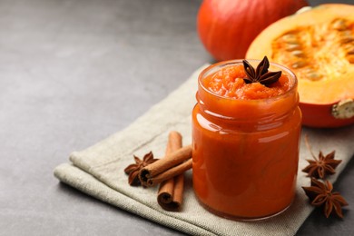 Photo of Jar of pumpkin jam, star anise, cinnamon and fresh pumpkin on grey table, closeup. Space for text