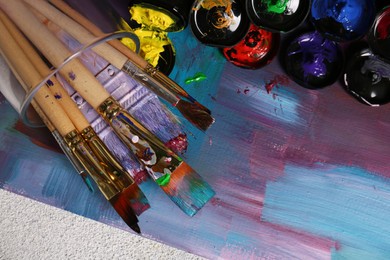 Photo of Canvas with colorful abstract painting and different brushes, top view