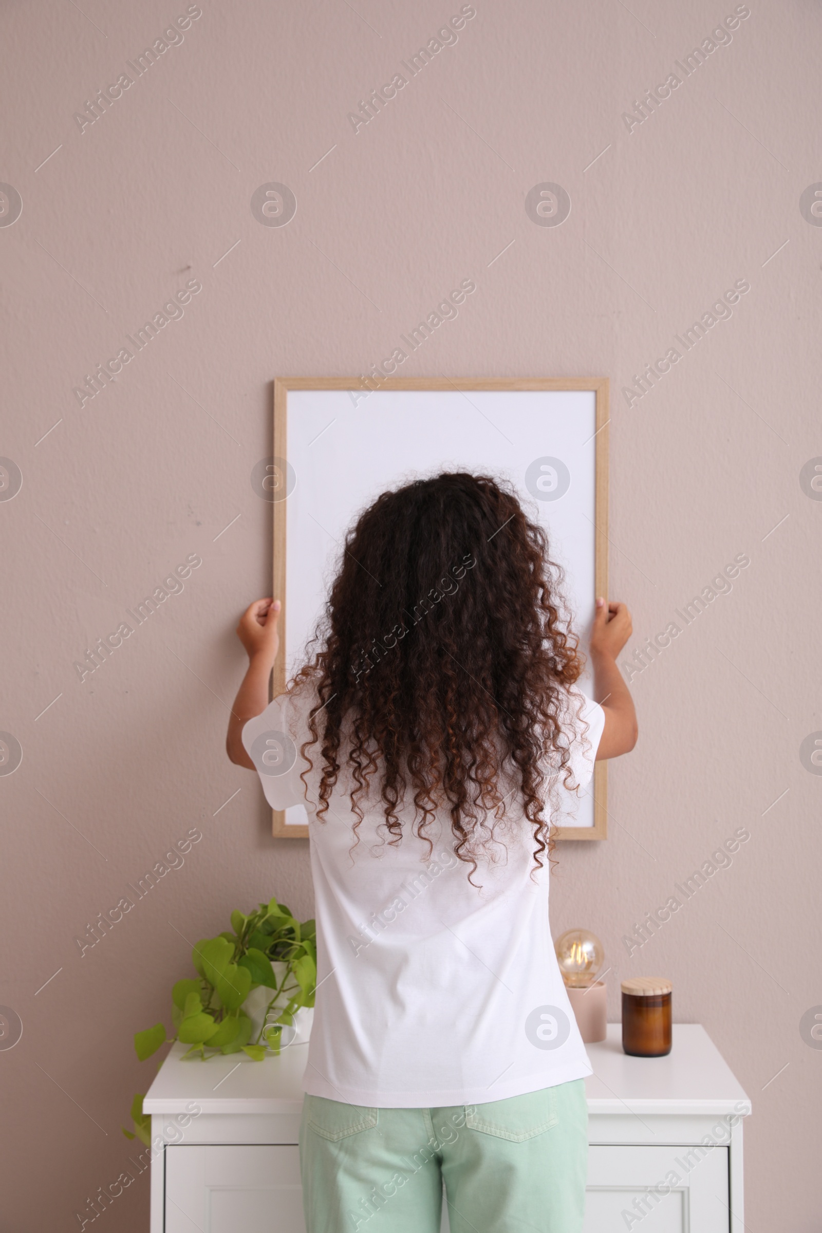 Photo of Woman hanging frame on pale rose wall over chest of drawers in room, back view