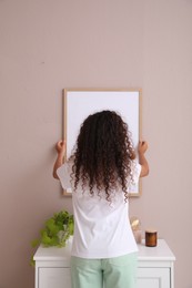 Photo of Woman hanging frame on pale rose wall over chest of drawers in room, back view