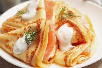 Photo of Delicious thin pancakes with salmon and sour cream on plate, closeup