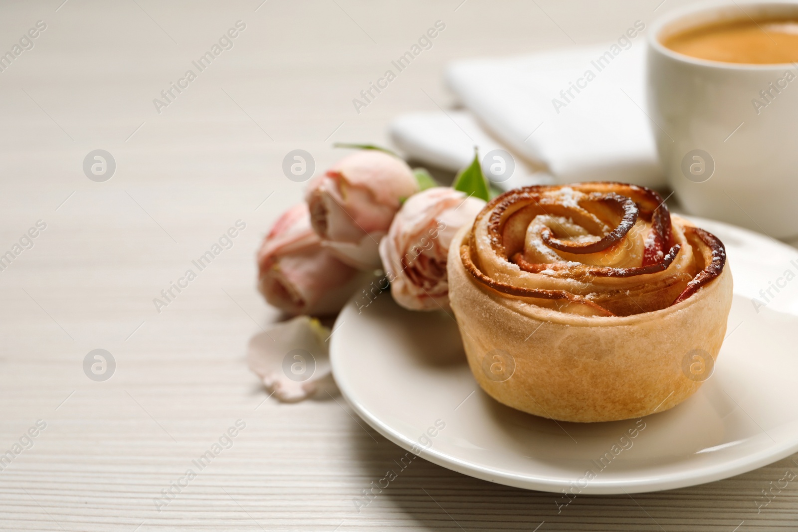 Photo of Freshly baked apple rose, cup of coffee and beautiful flowers on white wooden table, space for text