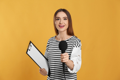 Young female journalist with microphone and clipboard on yellow background