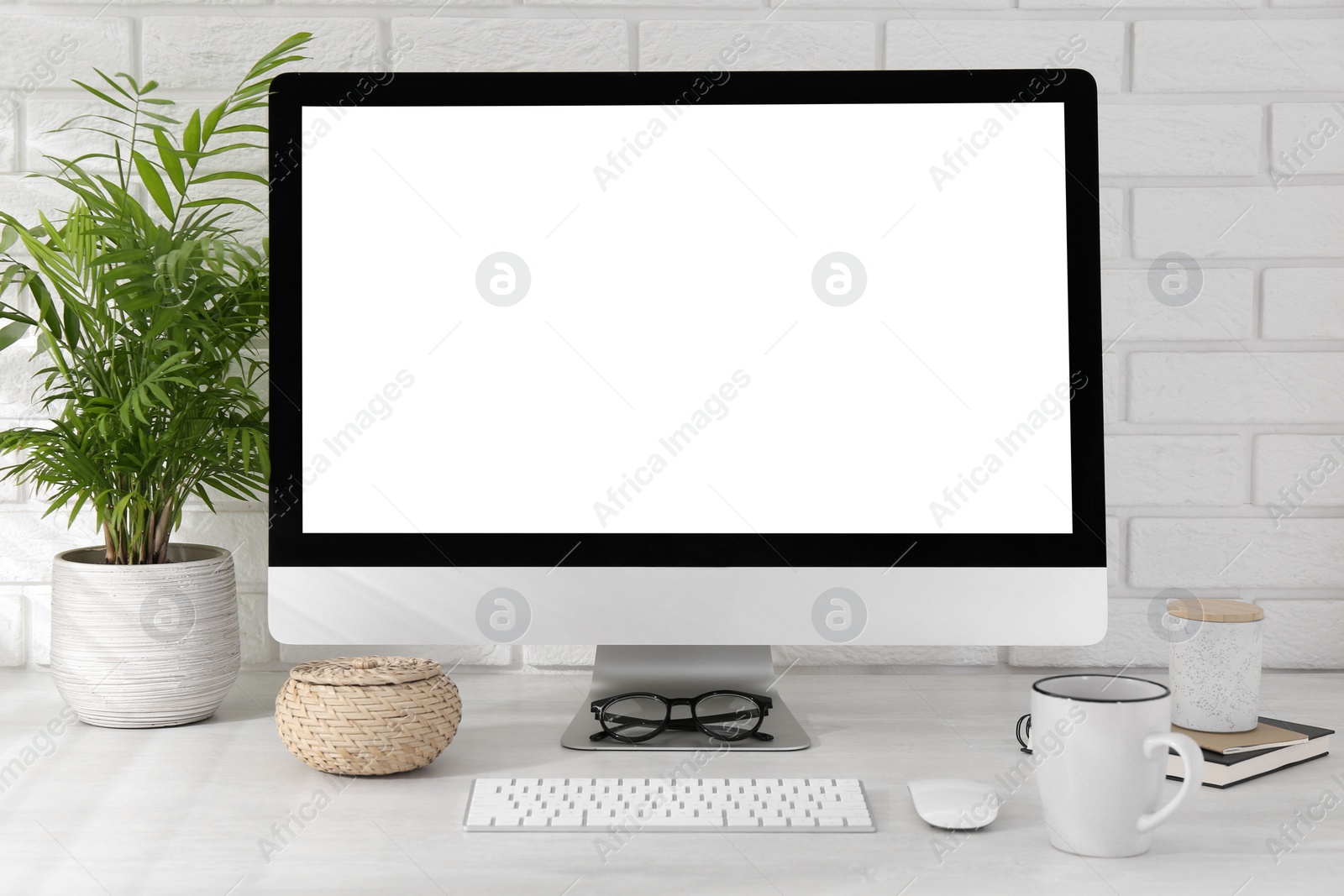 Photo of Office workplace with computer, cup, glasses, stationery and houseplant on light table near white brick wall