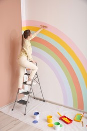 Young woman painting rainbow on white wall indoors
