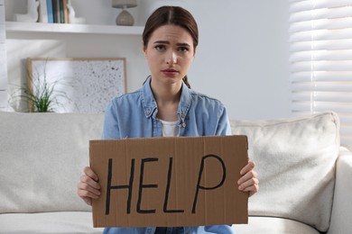 Unhappy young woman with HELP sign on sofa indoors