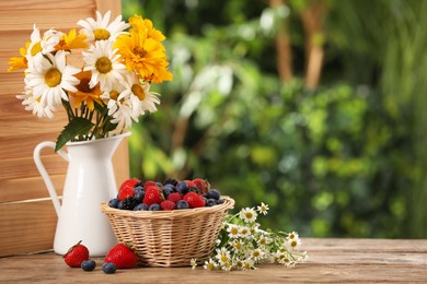 Photo of Wicker bowl with different fresh ripe berries and beautiful flowers on wooden table outdoors, space for text