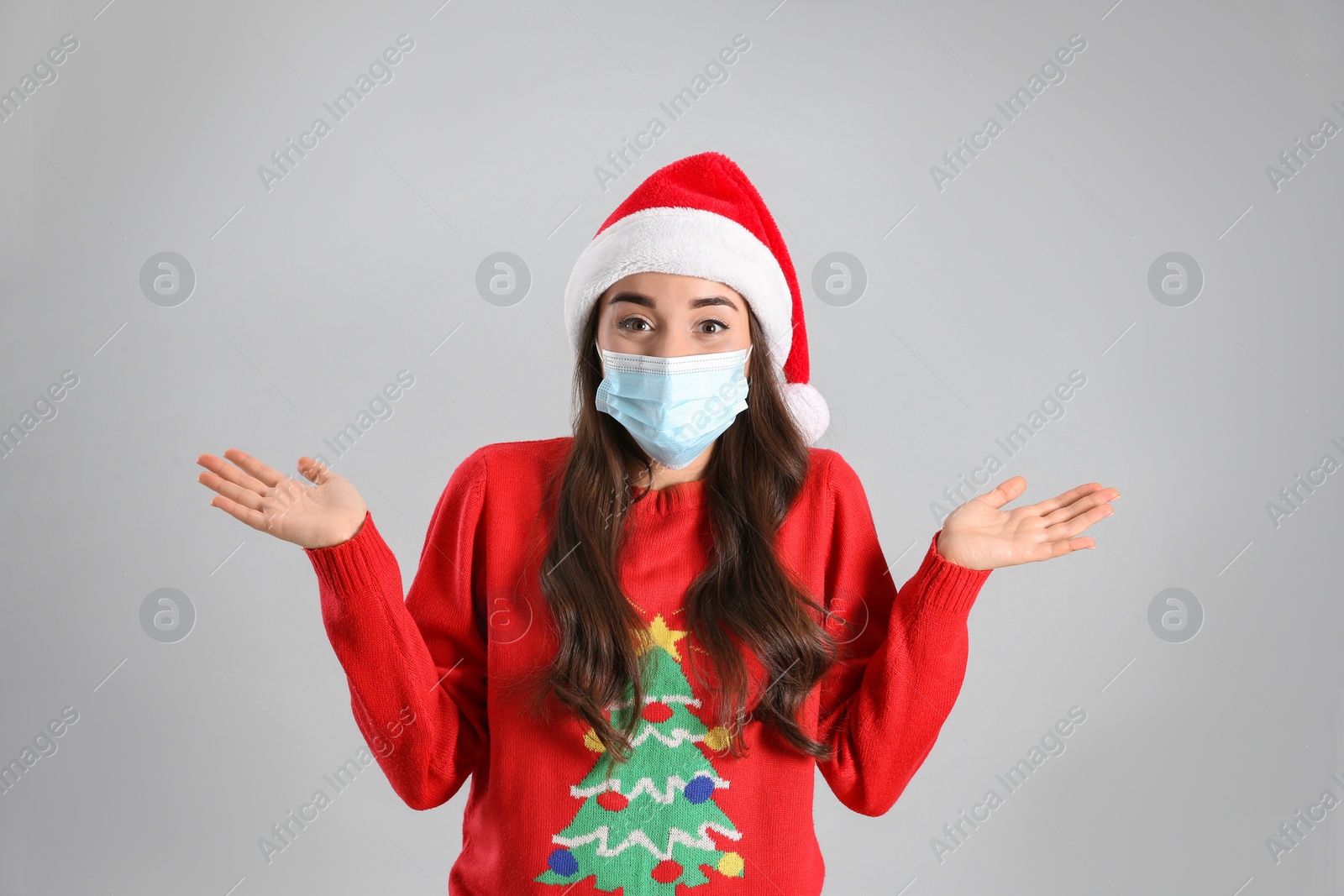 Photo of Beautiful emotional woman wearing Santa Claus hat and medical mask on light grey background