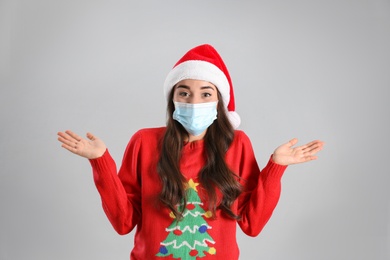 Beautiful emotional woman wearing Santa Claus hat and medical mask on light grey background