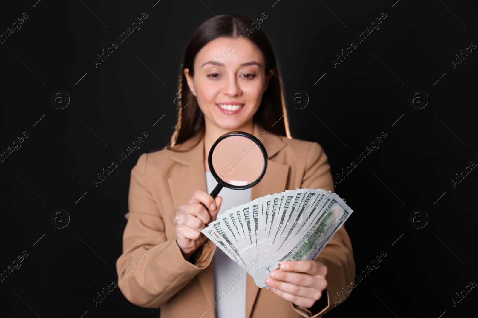 Photo of Expert authenticating 100 dollar banknotes with magnifying glass against black background, focus on hand. Fake money concept