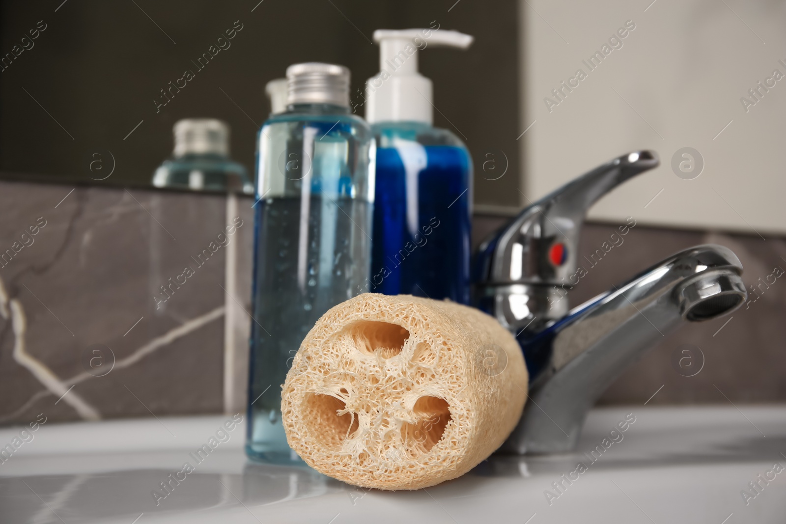 Photo of Natural loofah sponge and shower gel bottles on washbasin in bathroom, closeup