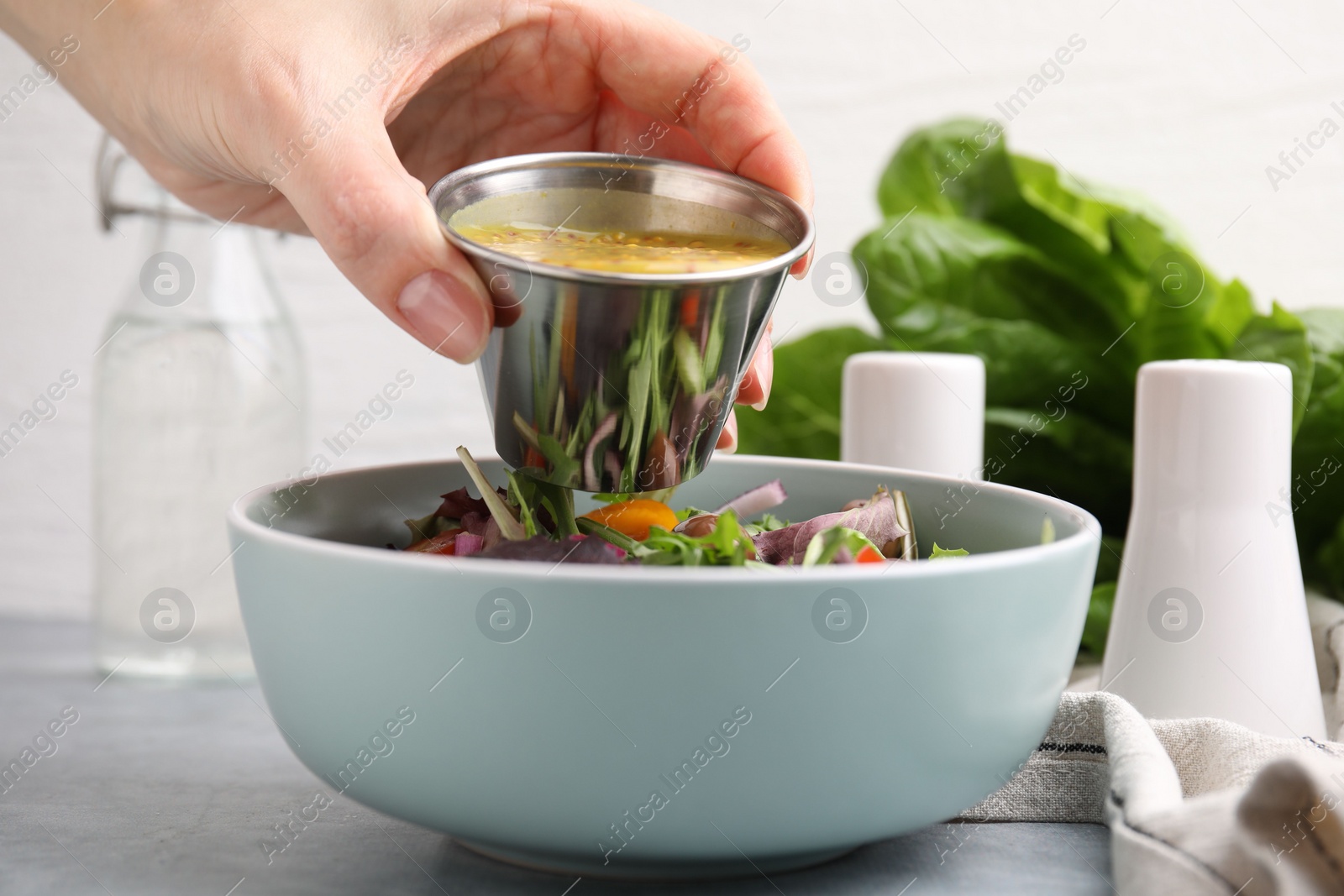 Photo of Woman pouring tasty vinegar based sauce (Vinaigrette) into bowl with salad at grey table, closeup