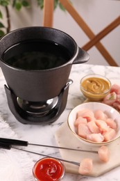 Fondue pot with oil, forks, raw meat pieces, sauce and mustard on white marble table