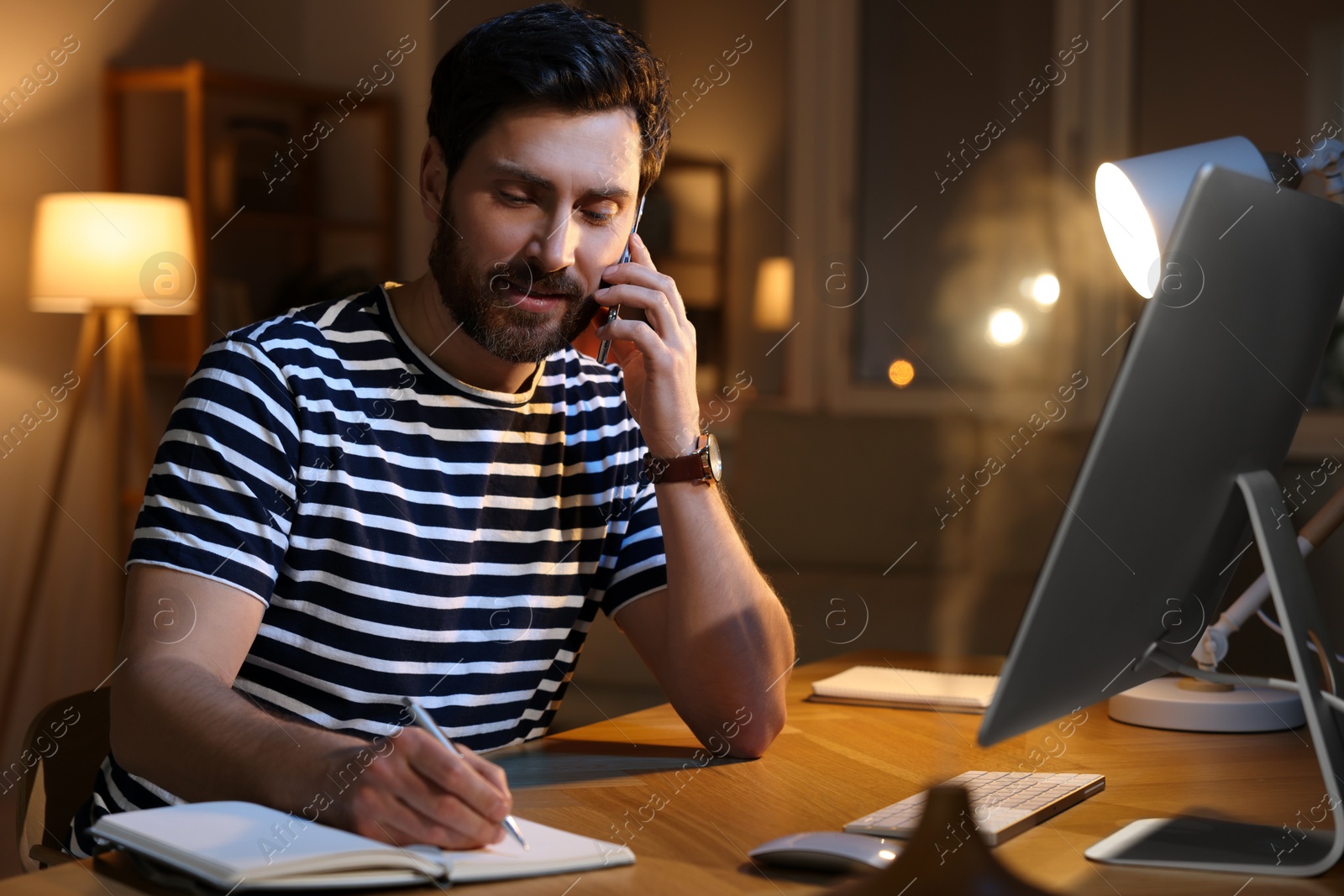 Photo of Home workplace. Man taking notes while talking on smartphone at wooden desk in room at night