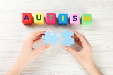 Photo of Woman holding puzzle pieces near cubes with word Autism at white wooden table, top view