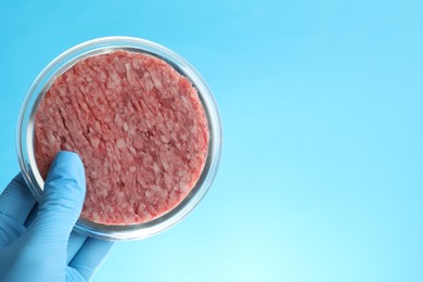 Photo of Scientist holding Petri dish with minced lab grown meat on light blue background, closeup. Space for text