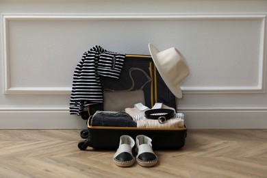 Open suitcase with folded clothes, accessories and shoes near white wall indoors