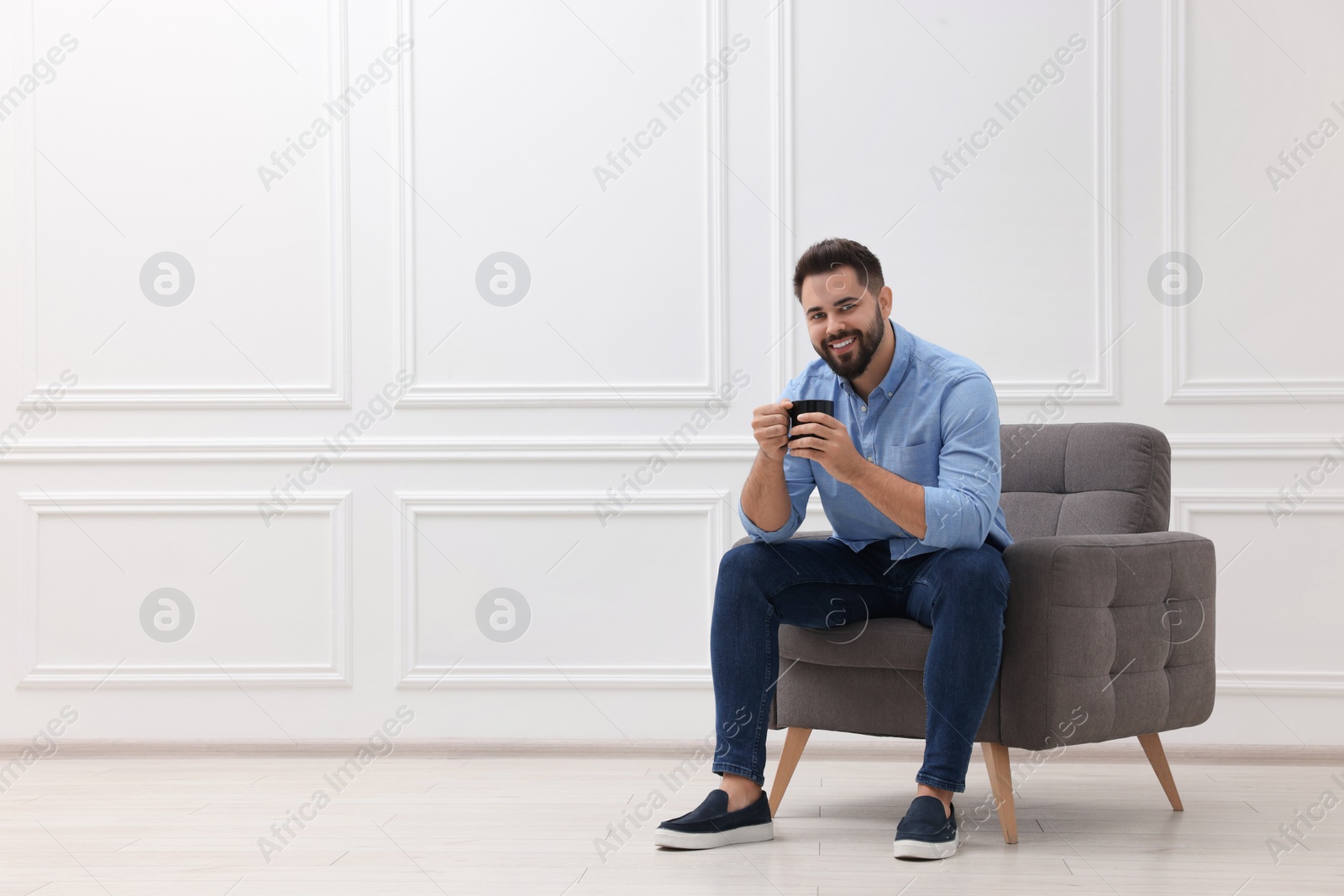 Photo of Handsome man with cup of drink sitting in armchair near white wall indoors, space for text