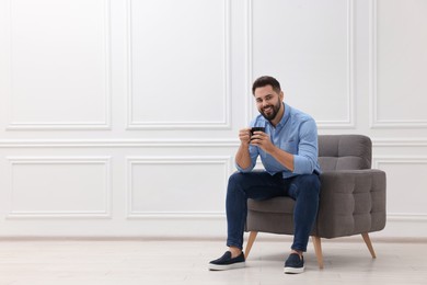 Photo of Handsome man with cup of drink sitting in armchair near white wall indoors, space for text