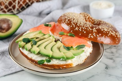 Photo of Delicious bagel with cream cheese, salmon, avocado and microgreens on white marble table, closeup