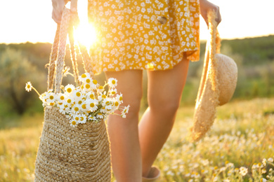 Photo of Woman with straw hat and handbag full of chamomiles walking in meadow, closeup
