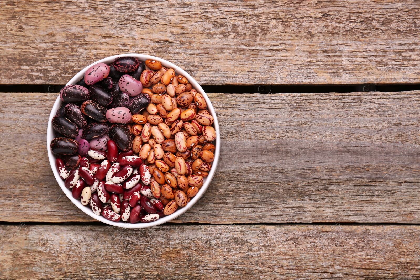 Photo of Different kinds of dry kidney beans in bowl on wooden table, top view. Space for text