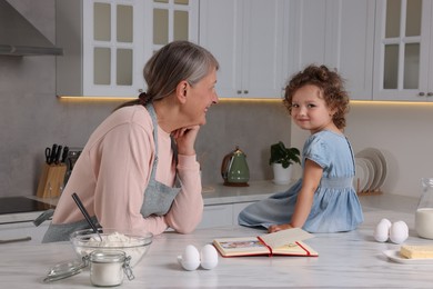 Photo of Cute little girl with her granny cooking by recipe book in kitchen