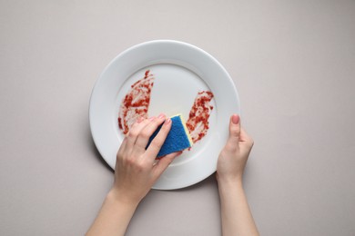 Woman washing dirty plate with sponge on light grey background, top view