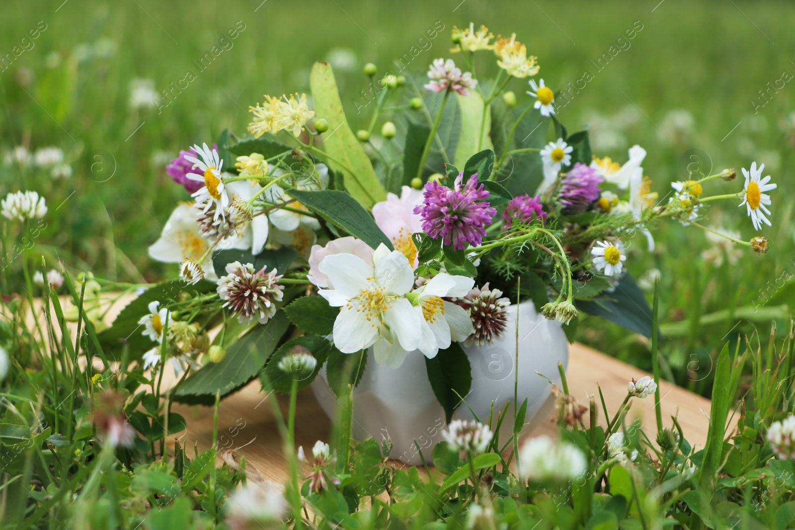 Photo of Ceramic mortar with different wildflowers and herbs on wooden board in meadow