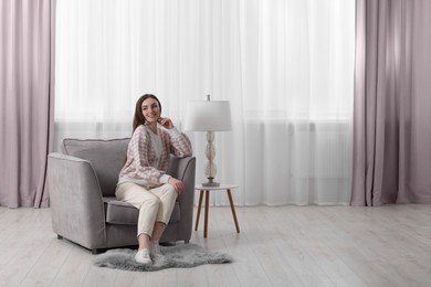 Woman sitting on armchair near lamp and window with stylish curtains at home. Space for text
