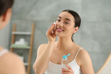 Beautiful woman removing makeup with cotton pad near mirror indoors