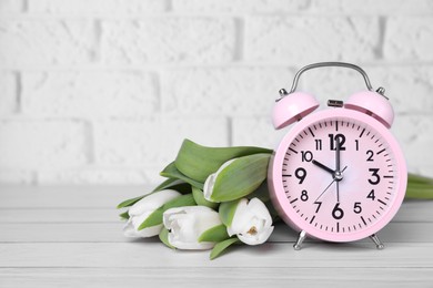 Photo of Pink alarm clock and beautiful tulips on white wooden table against brick wall, space for text. Spring time