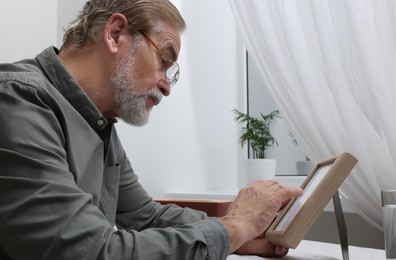 Photo of Upset senior man with photo frame at table in room. Loneliness concept