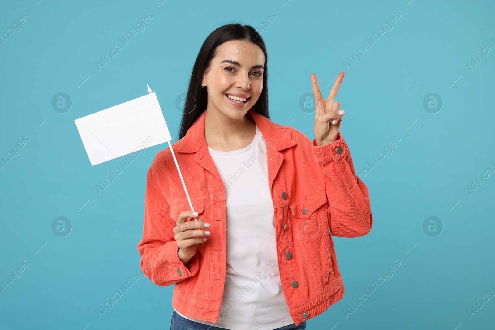 Photo of Happy young woman with blank white flag showing V-sign on light blue background. Mockup for design