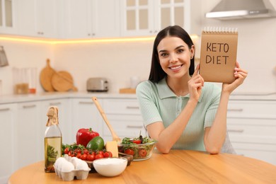 Photo of Happy woman holding notebook with words Keto Diet near different products in kitchen