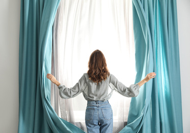 Photo of Woman opening window curtains at home in morning, back view