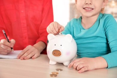 Little girl putting coin into piggy bank and her mother at table, closeup. Saving money