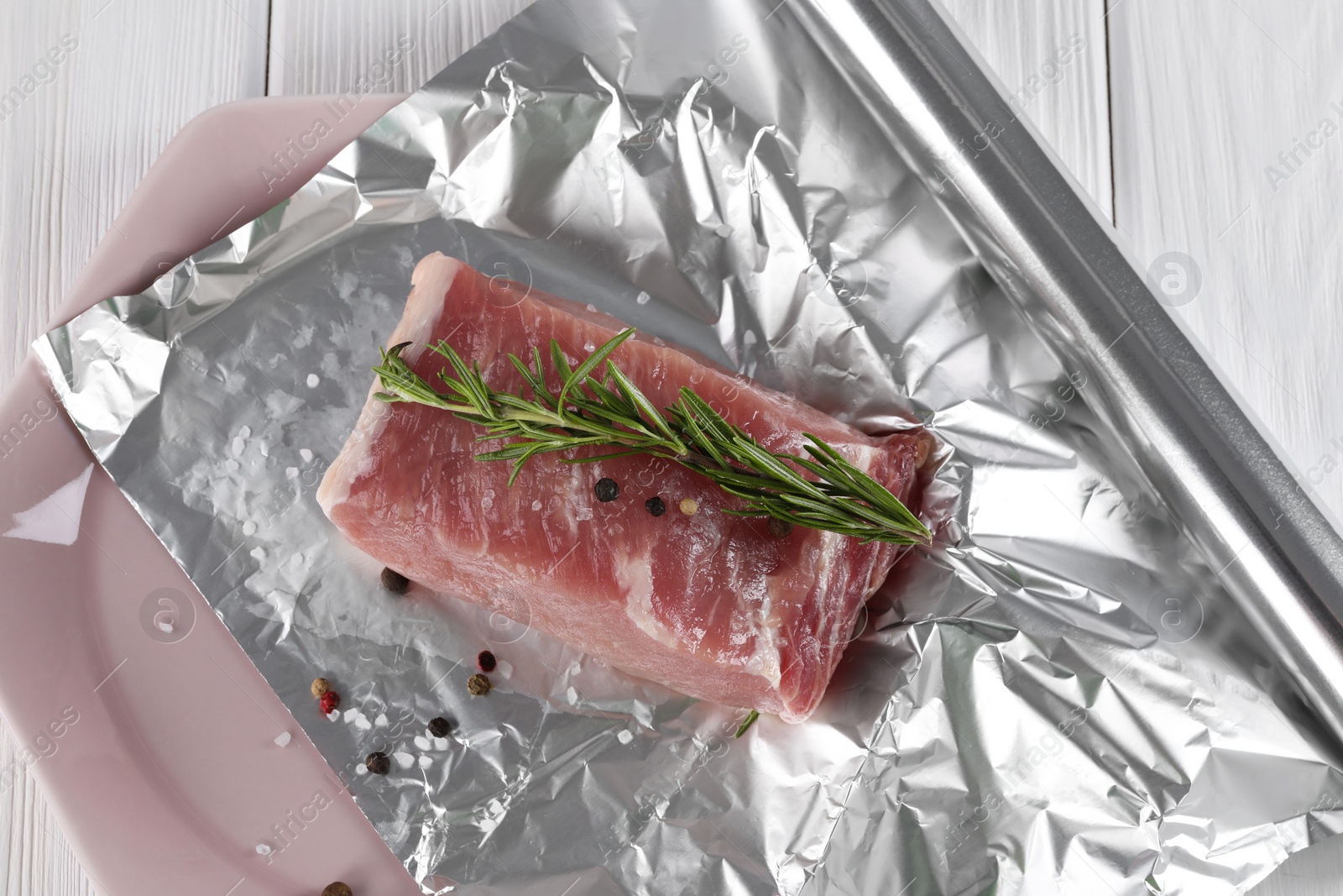 Photo of Aluminum foil with raw meat, rosemary and spices on white wooden table, top view