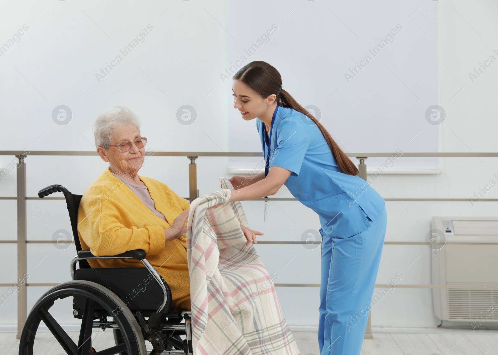 Photo of Nurse covering senior woman in wheelchair with plaid at hospital. Medical assisting