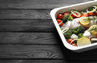 Photo of Raw fish with vegetables and lemon in baking dish on black wooden table. Space for text