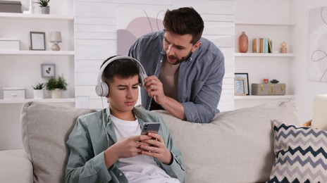 Photo of Father scolding his son while he listening music in headphones at home. Teenager problems