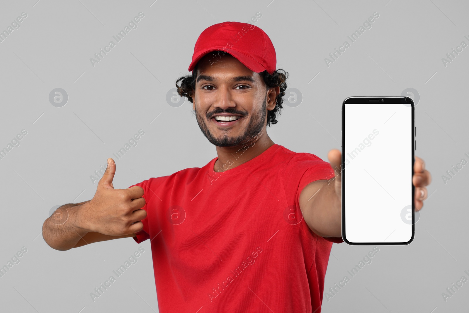 Image of Happy courier holding smartphone with empty screen and showing thumbs up on light grey background